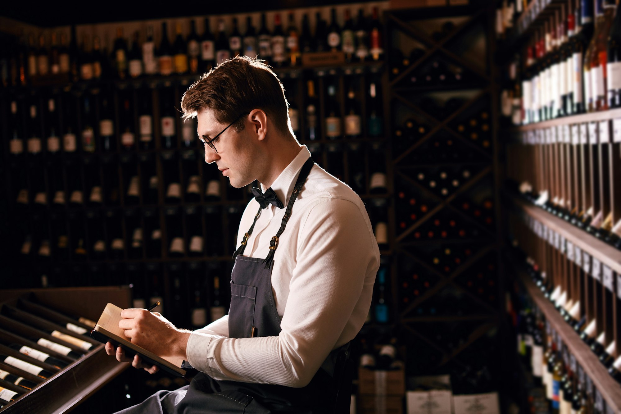 Male wine steward looks at bottles of wine and writing in notebook at wine store Male wine steward looks at bottles of wine and writing in notebook at wine store